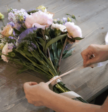 Händer binder ett band runt en bukett med färgglada blommor på ett träbord.