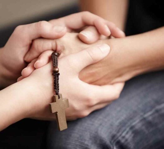 Två händer håller varandra, en håller ett radband med ett kors.