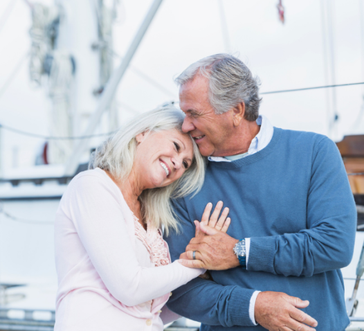 An older couple embraces affectionately on a boat, with the woman leaning her head on the man's shoulder and both smiling warmly.